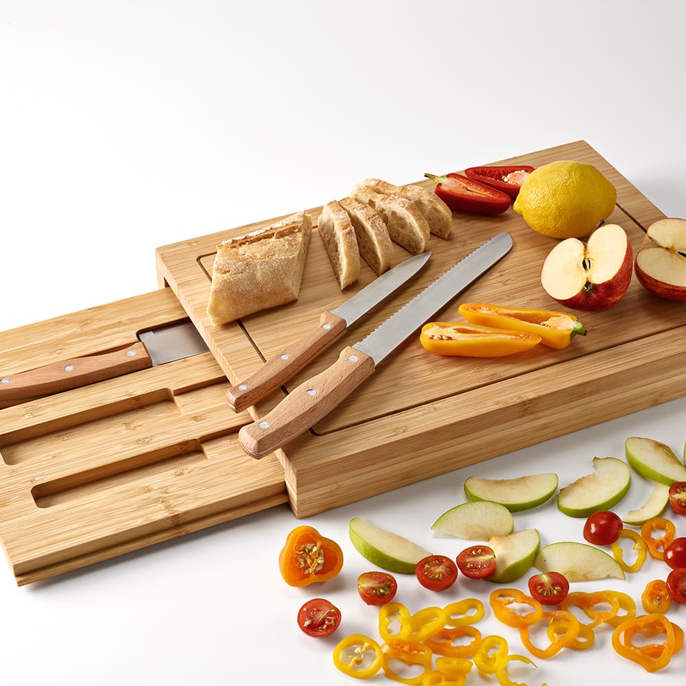 A bamboo cutting board set with kitchen knives, sliced bread, lemon, and assorted fresh vegetables and fruits.