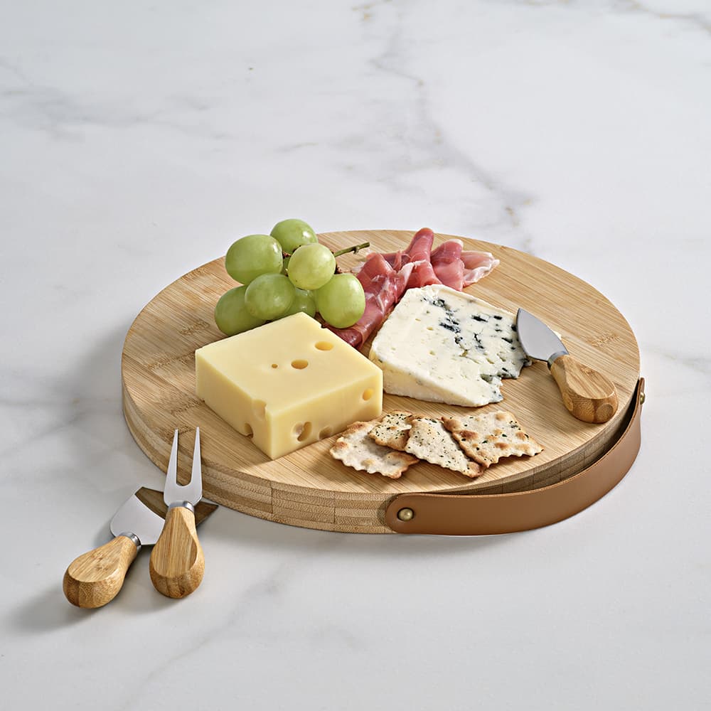 A round bamboo cheese board featuring various cheeses, crackers, grapes, and utensils with wooden handles.