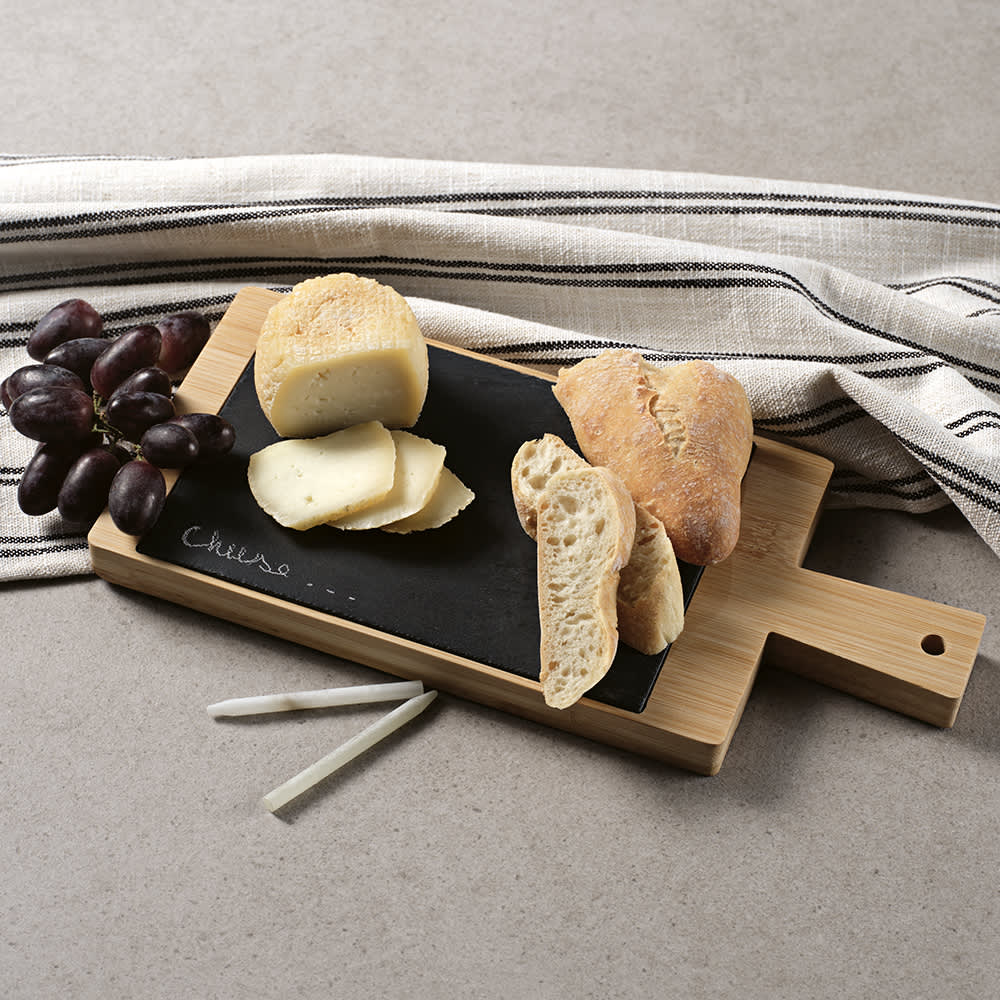 A bamboo and slate serving board with a handle, featuring cheese, bread, grapes, and chalk.