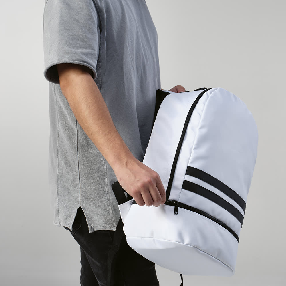 A white backpack with reflective front strips, held by a person wearing a grey shirt.