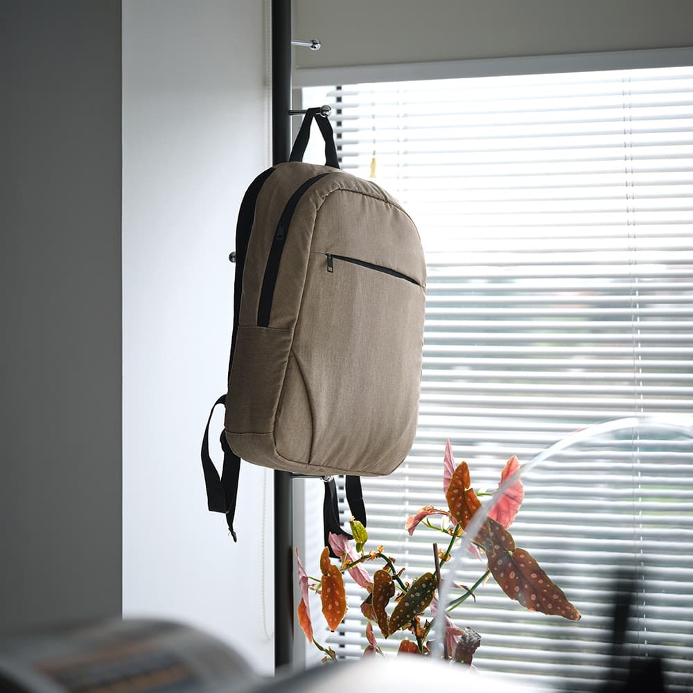 A tan backpack with a front pocket, designed to fit a 17" laptop, hanging by a window.