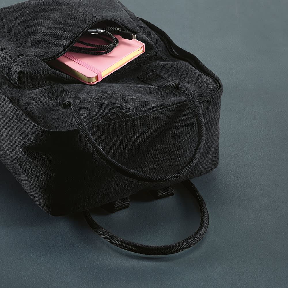 A black backpack with rPET handles sits on a surface, partially open with a pink notebook visible inside.