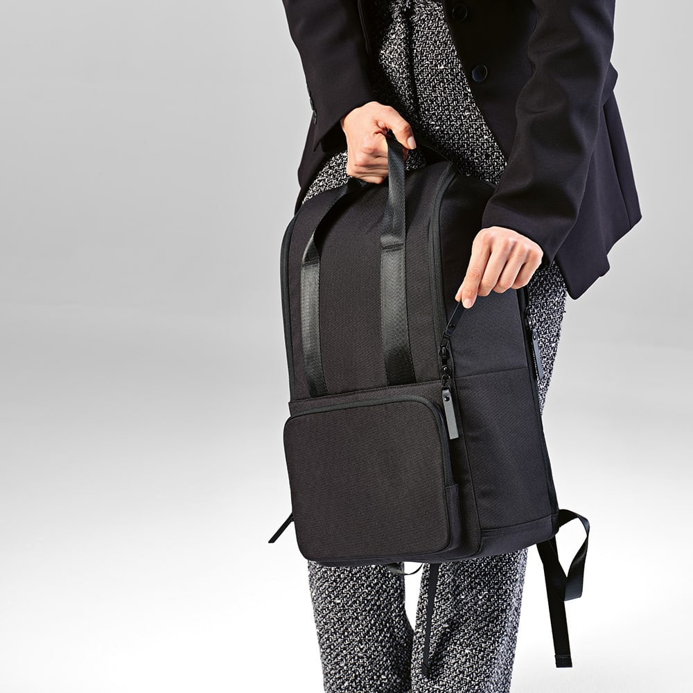 A black backpack with a front pocket, designed to fit a 15.6" laptop, held by a person in formal attire.