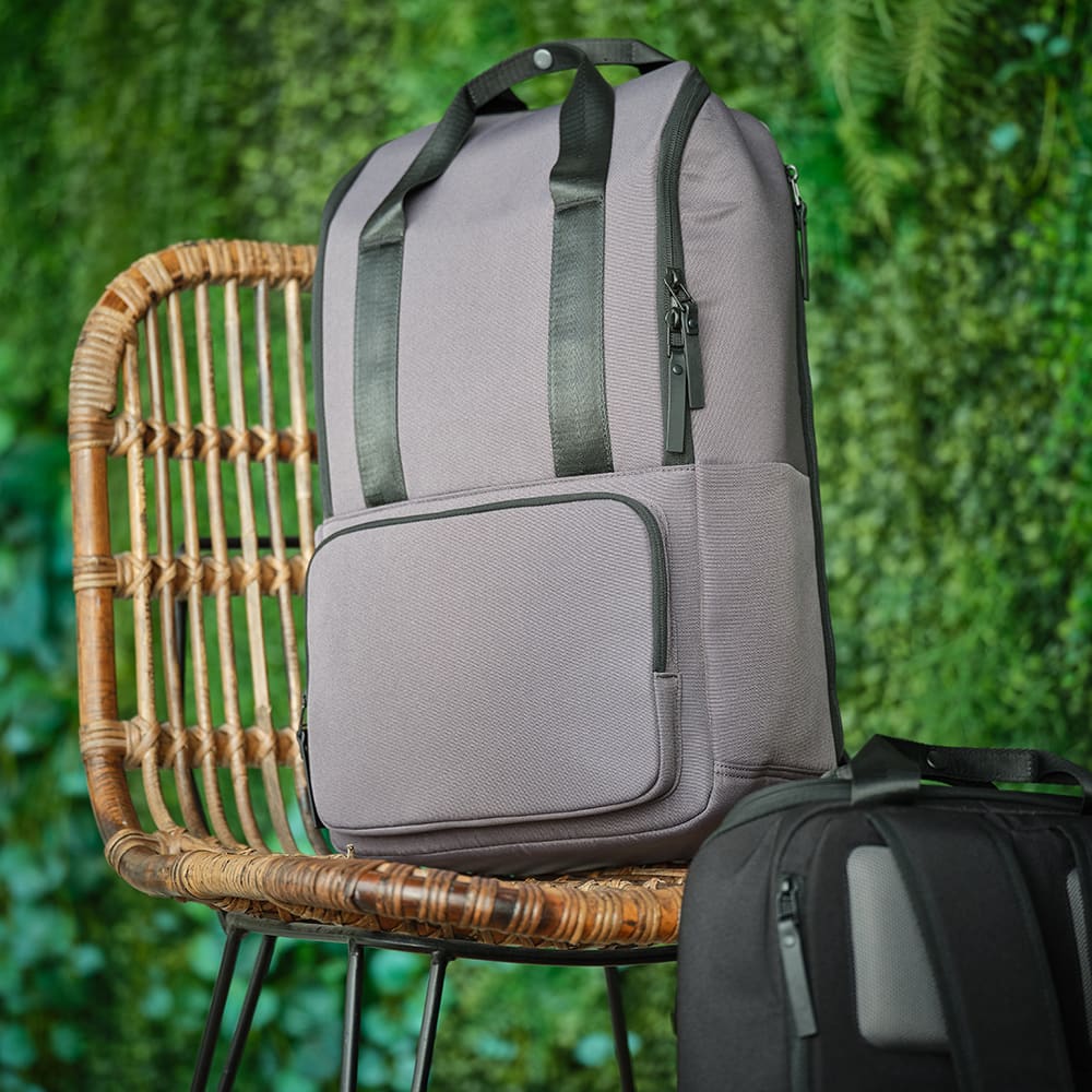 A grey backpack with two black straps, featuring a front pocket, displayed on a chair against a green background.