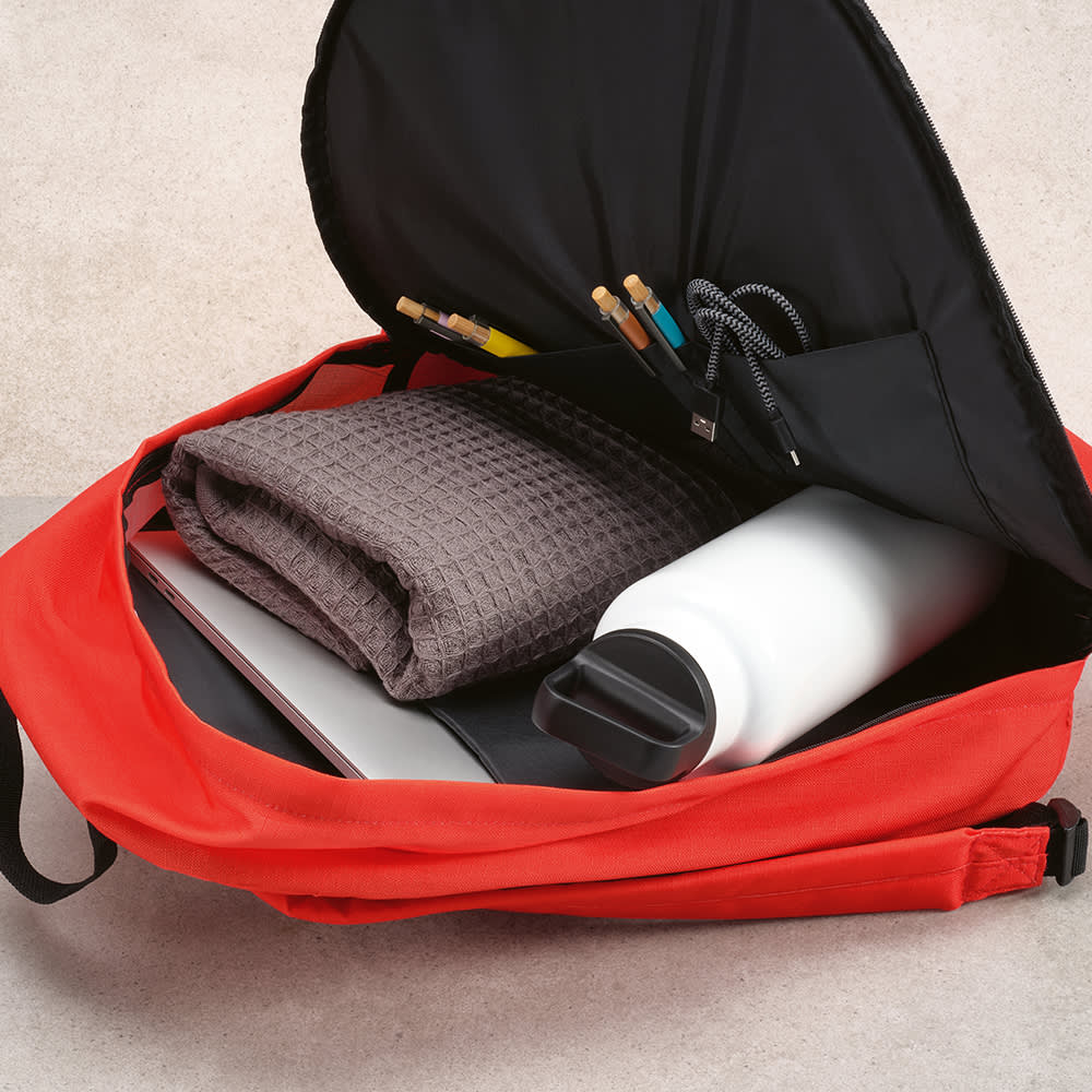 A red backpack with a black interior, containing a grey blanket, laptop, drink bottle, and stationery items.