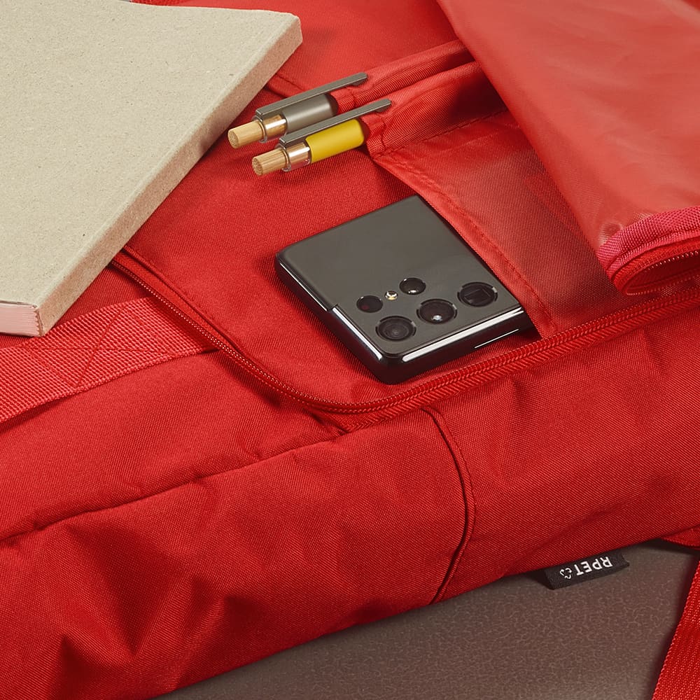 A red backpack featuring a roll top and buckle closure, with a phone, notebook, and stationery inside.