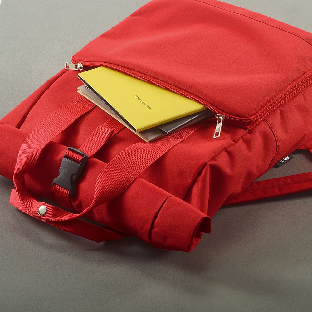 A red backpack with a roll top and buckle closure, featuring exposed zippers and documents inside.