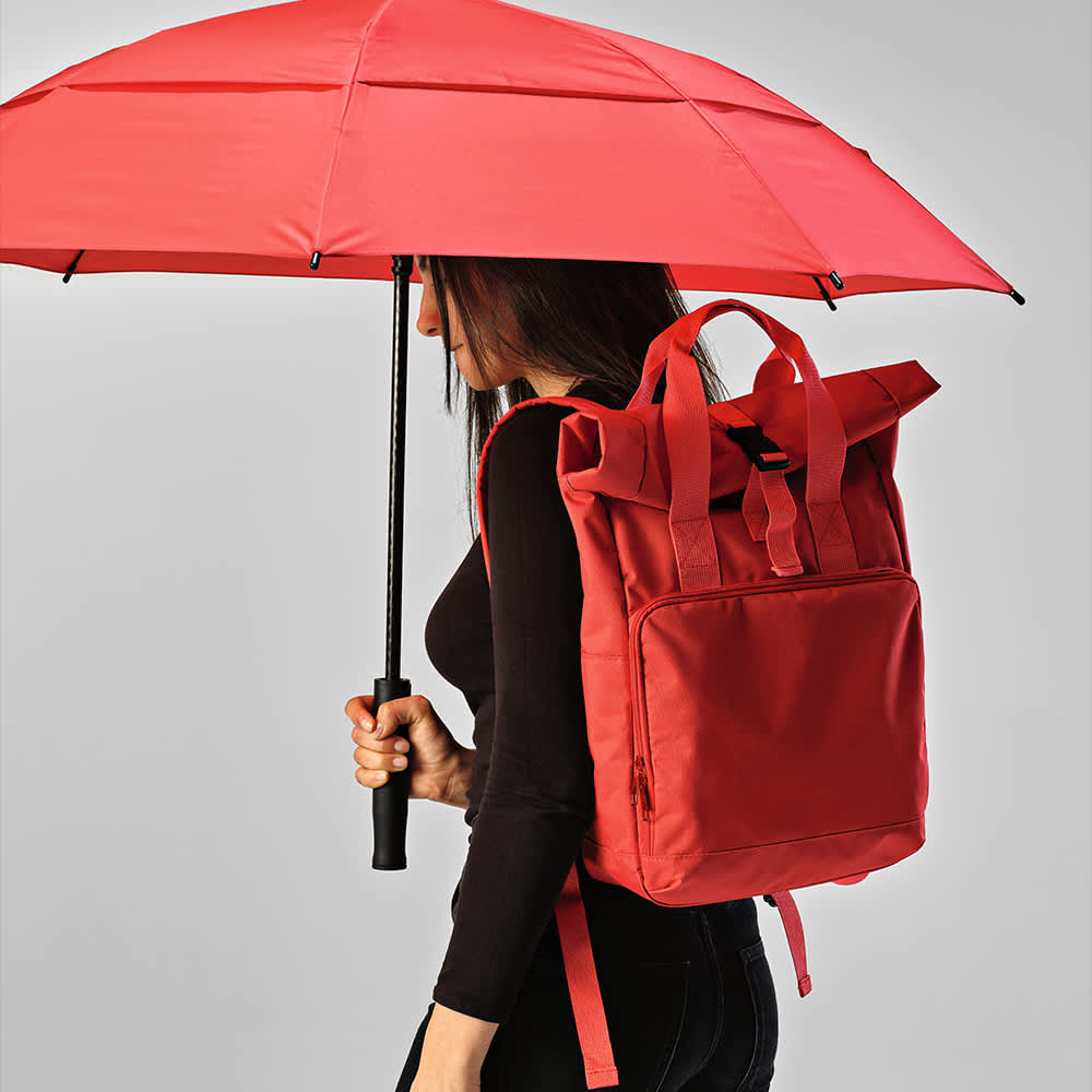 A red roll-top backpack with buckle closure, carried by a person holding a matching red umbrella. The backpack has a logo.