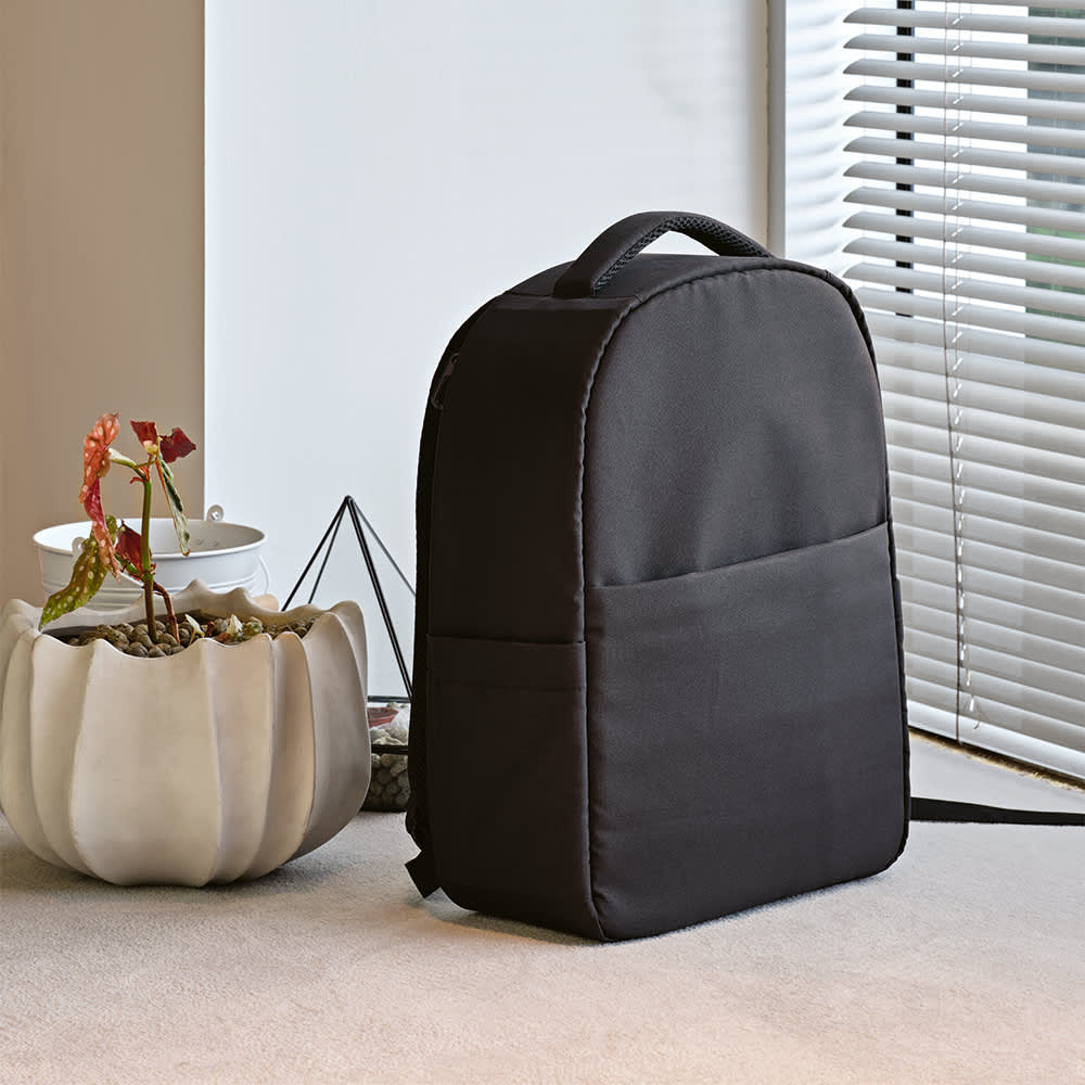 A black backpack with a front pocket, designed to fit a 17" laptop, resting near a plant and window.