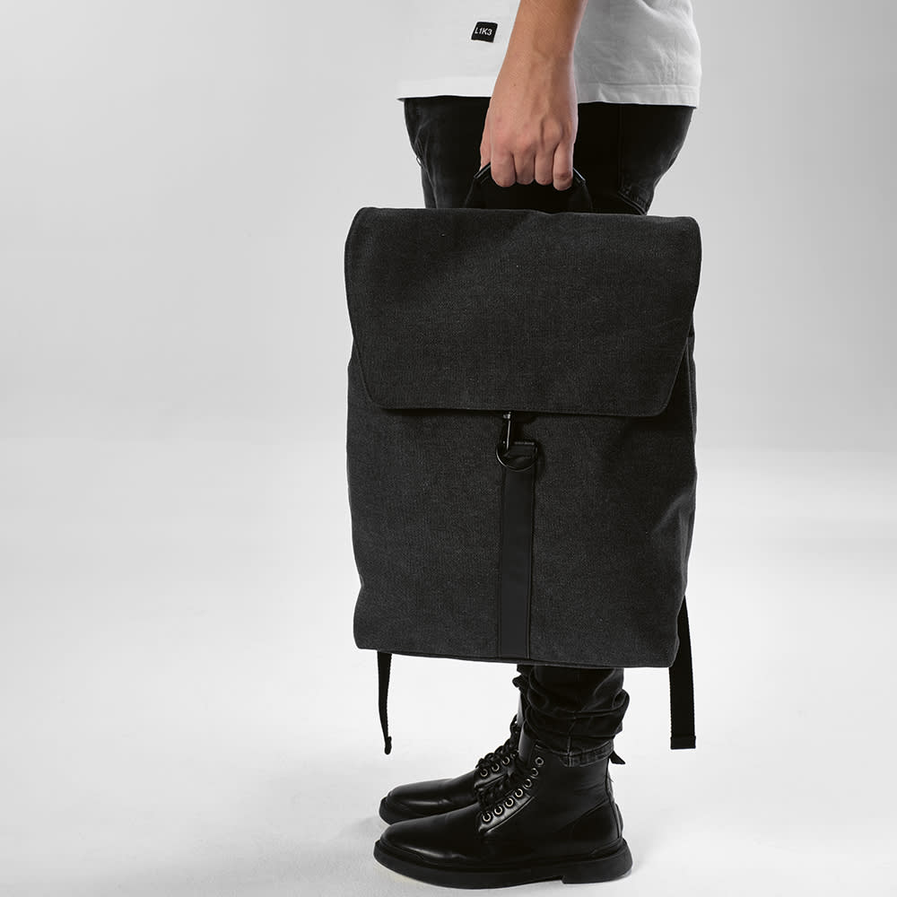 A black backpack with a hook closure, designed to fit a 17" laptop, held by a person wearing dark pants and boots.
