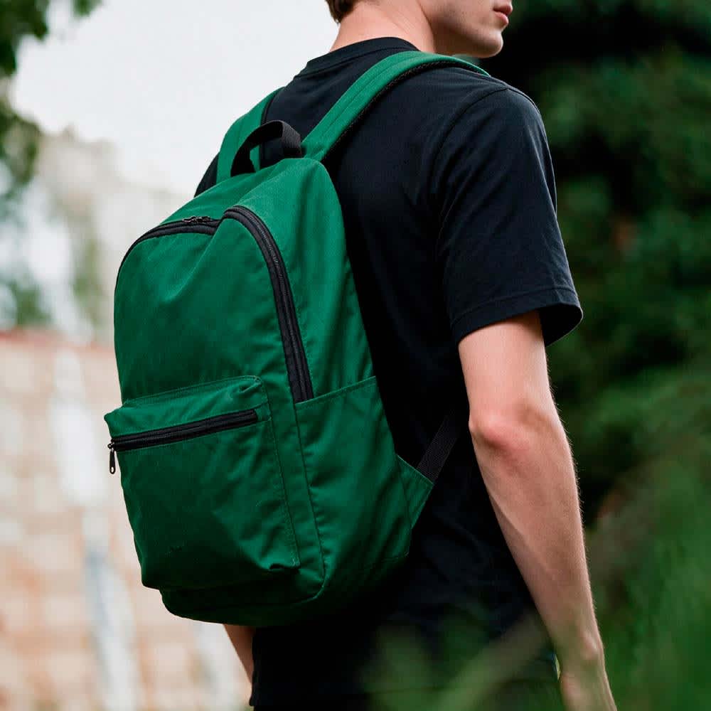 A green backpack with a front zipper pocket, worn by a person in a black shirt, against a blurred outdoor background.