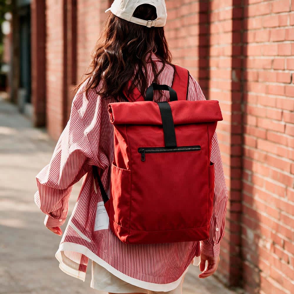 A red roll-top backpack with a velcro closure, worn on the back, against a brick wall background.