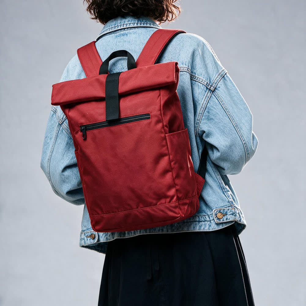 A red roll-top backpack with a velcro closure and a side pocket, worn by a person in a denim jacket.