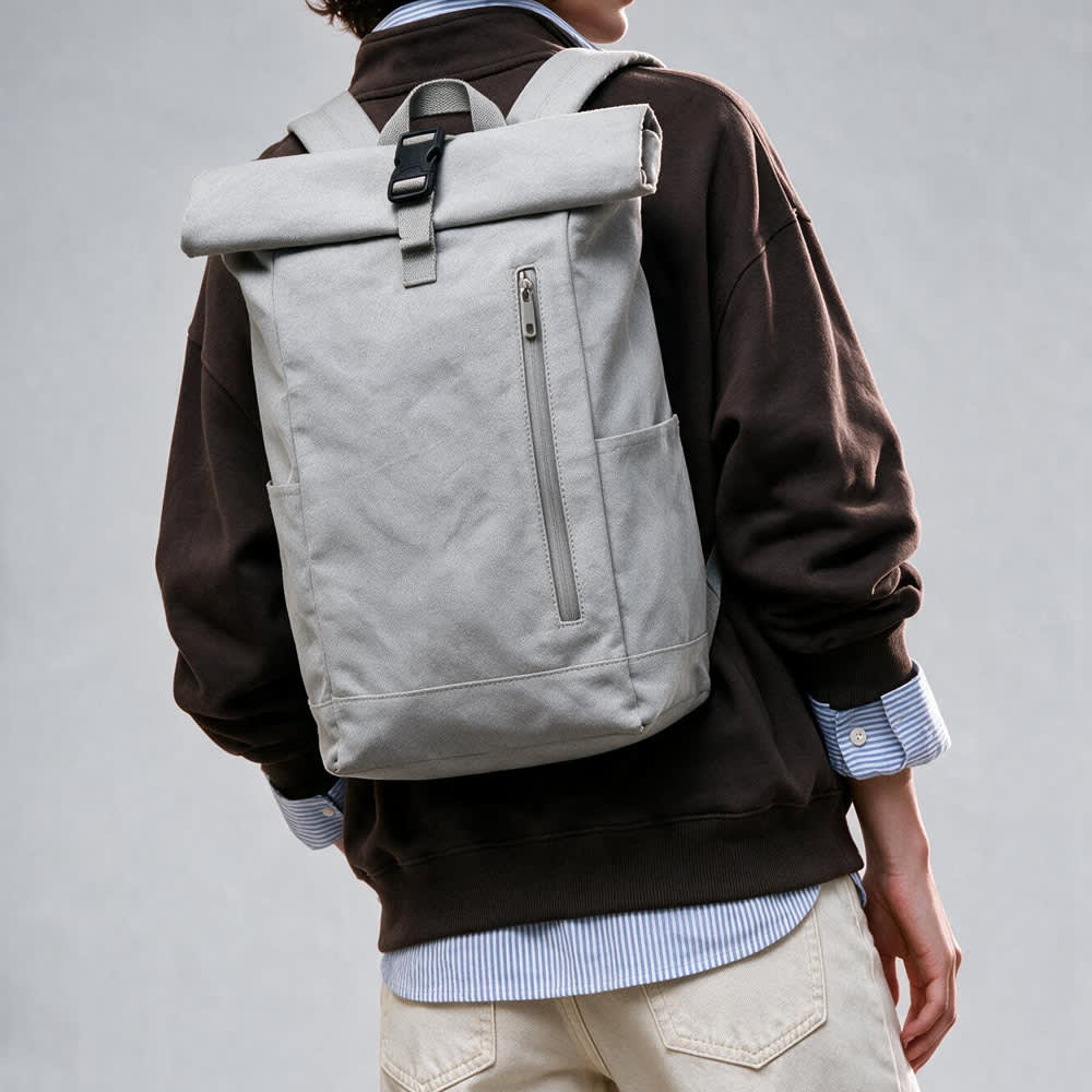 A grey roll-top backpack with a buckle closure and a side zip pocket, worn on a person's back.