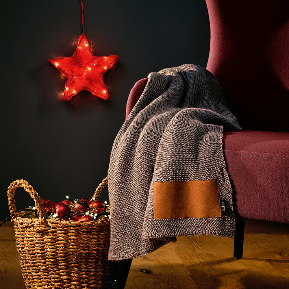 A grey blanket made from rPET and wool blend drapes over a burgundy chair, with a brown customisable patch.