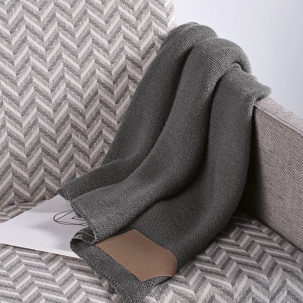 A grey wool blend blanket with a customisable patch draped over a textured sofa.