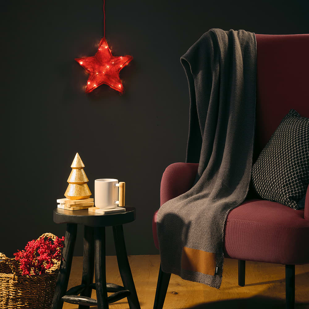 A grey wool blend blanket drapes over a maroon chair beside a small table with a white mug and decorative items.