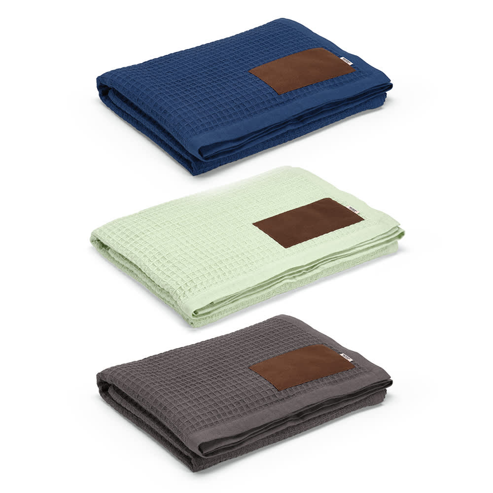 Three folded Giotto Blankets in navy blue, light green, and dark grey, each featuring a customisable brown patch.