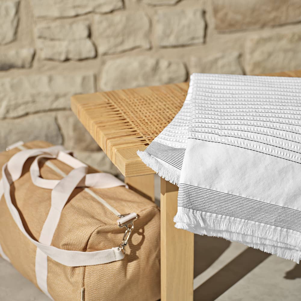 A beige duffle bag rests beside a white textured cotton blanket on a wooden table against a stone wall.