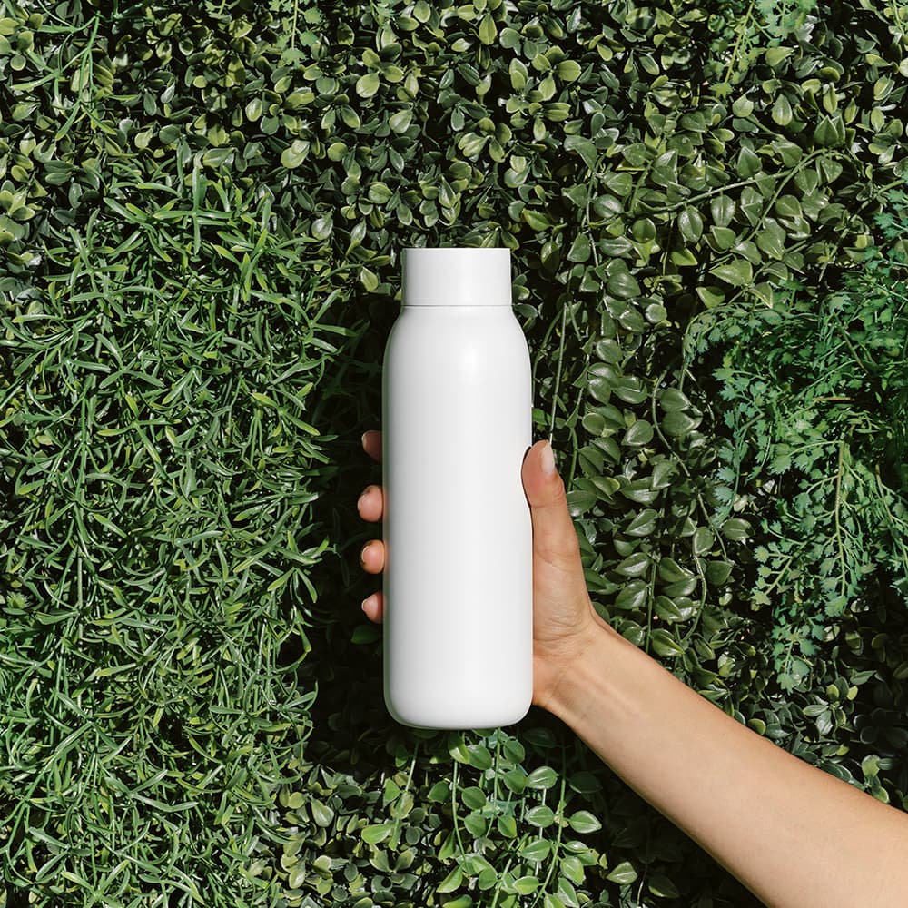 A drink bottle in white held against a background of green foliage, featuring a sleek design and slight curve.