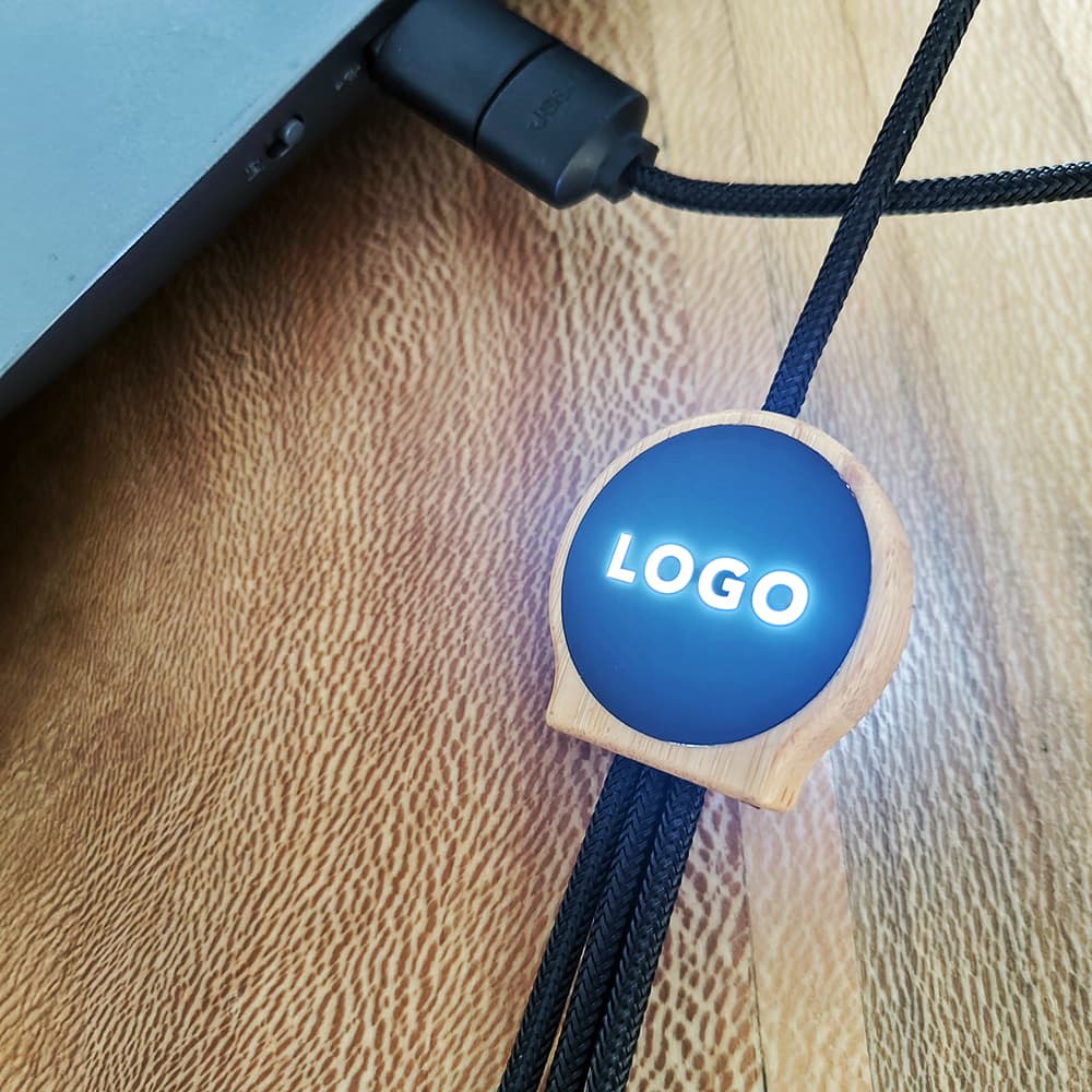 A black 5-in-1 cable featuring a bamboo and rPET holder with a backlit logo, placed near a laptop.