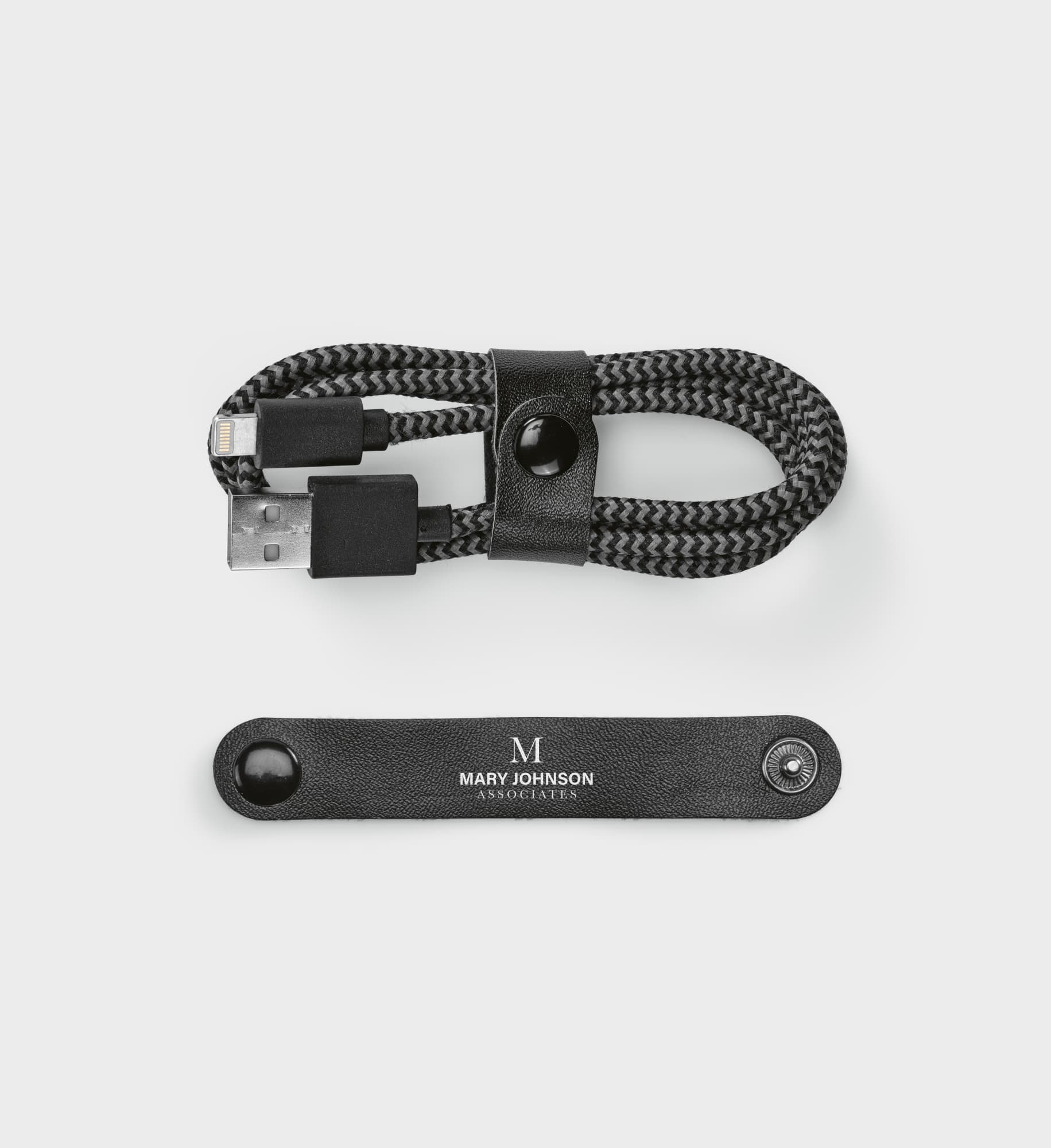 A black and grey braided USB-A to Lightning cable, secured with a leather strap featuring a logo.