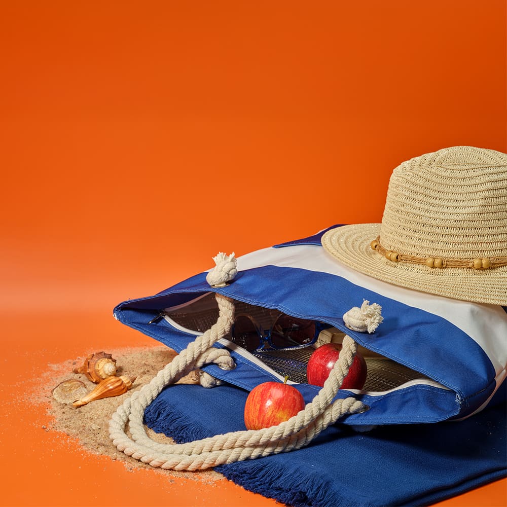 A blue and white cooler bag with rope handles, containing apples and a straw hat, set against an orange background.
