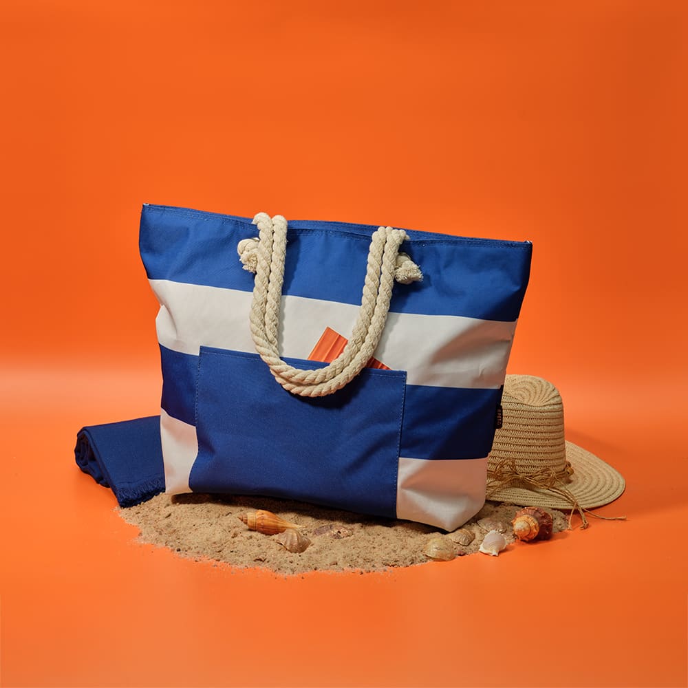 A large blue and white beach tote bag with rope handles, placed on sand alongside a straw hat and seashells.