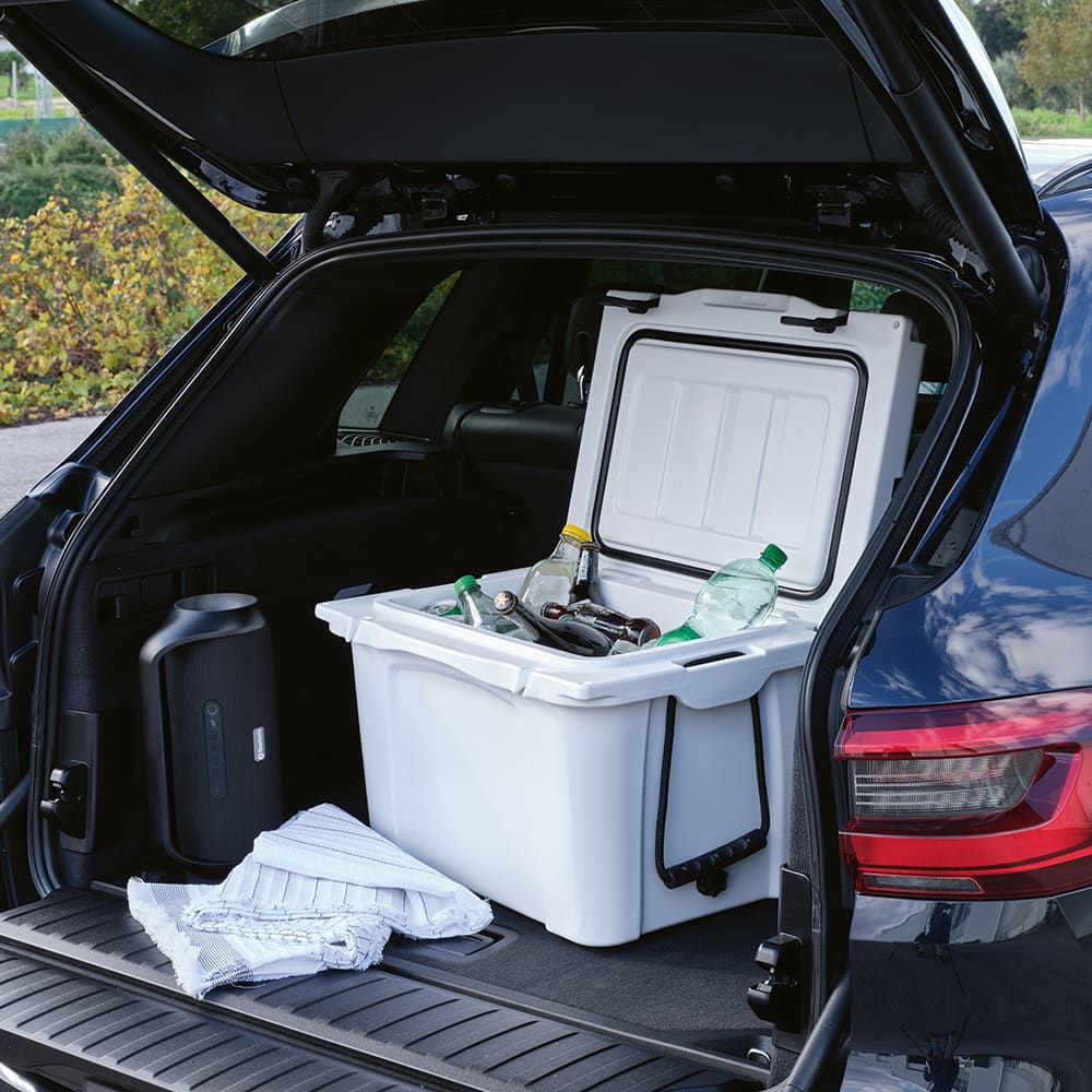 A white cooler sits in an open car boot, filled with various drinks and accompanied by towels. The car has a logo on it.