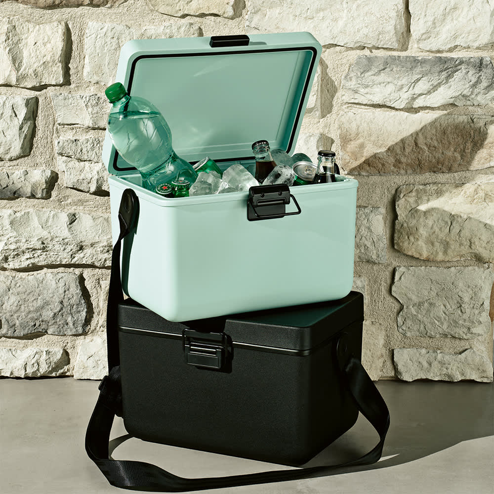 A drink cooler in pastel green, filled with bottles and ice, sits atop a black cooler against a stone wall.