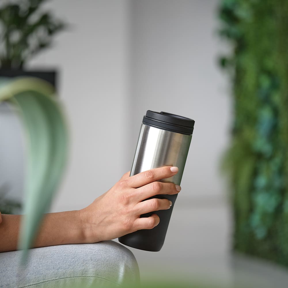 A drink bottle with a stainless steel body and a black lid, held by a hand against a green backdrop.
