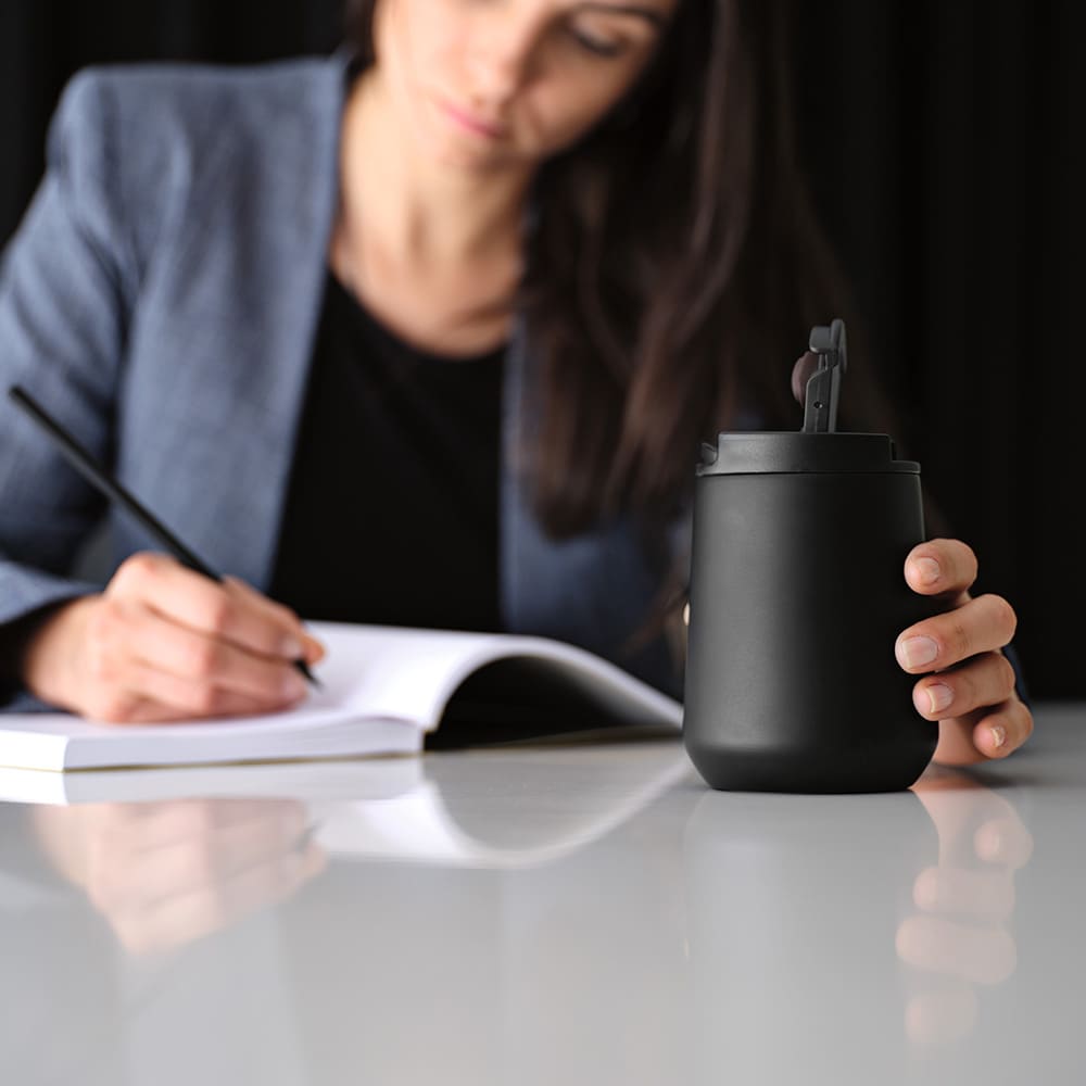 A drink bottle in matte black with a lid, resting on a table beside a person writing in a notebook.
