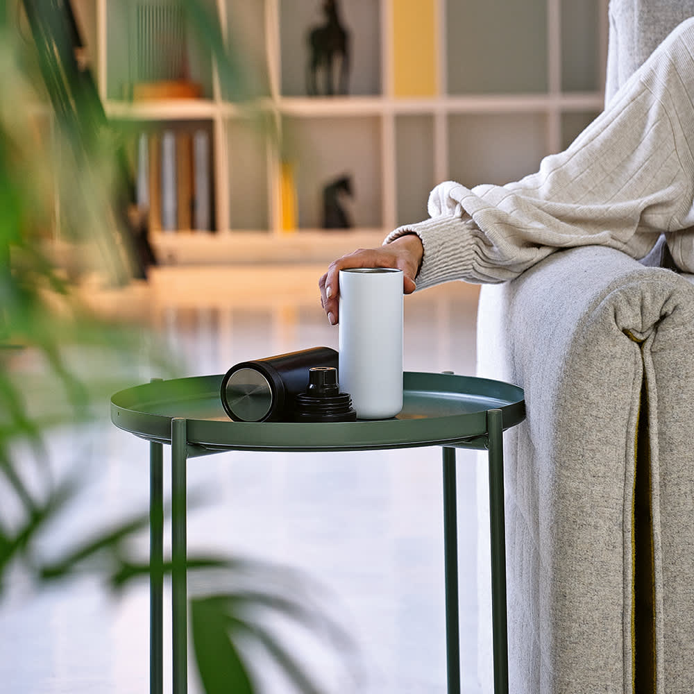 A drink bottle in white and black on a green side table, with a person’s hand resting nearby.