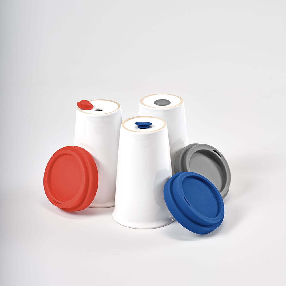 A set of ceramic travel cups in white with grey, blue, and red lids, featuring a logo on one cup.