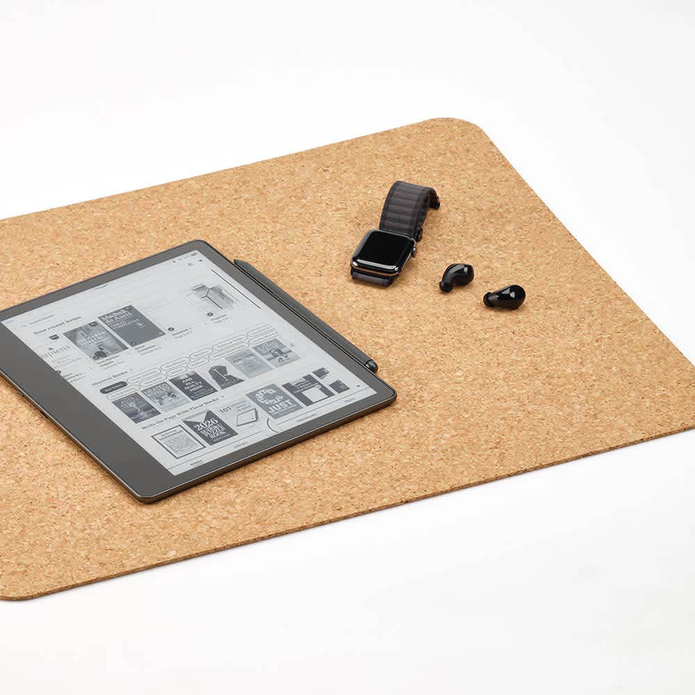 A cork desk mat in natural tones, featuring an e-reader, smartwatch, and earbuds on its surface.