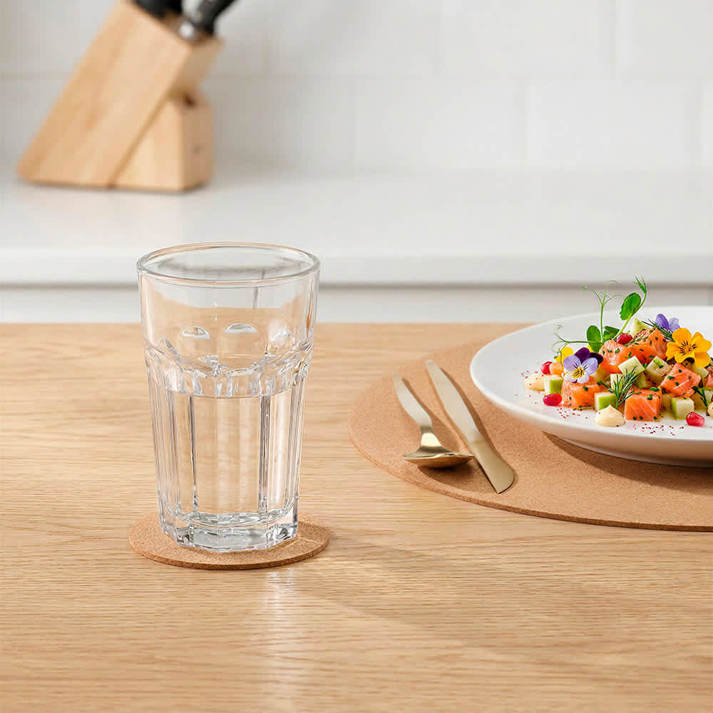 A drink glass rests on a round cork coaster on a wooden table, beside a plate of colourful salad garnished with flowers.