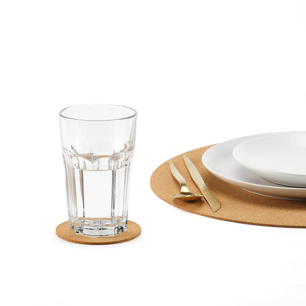 A clear drink glass sits on a round cork coaster, accompanied by white plates and gold utensils on a white surface.