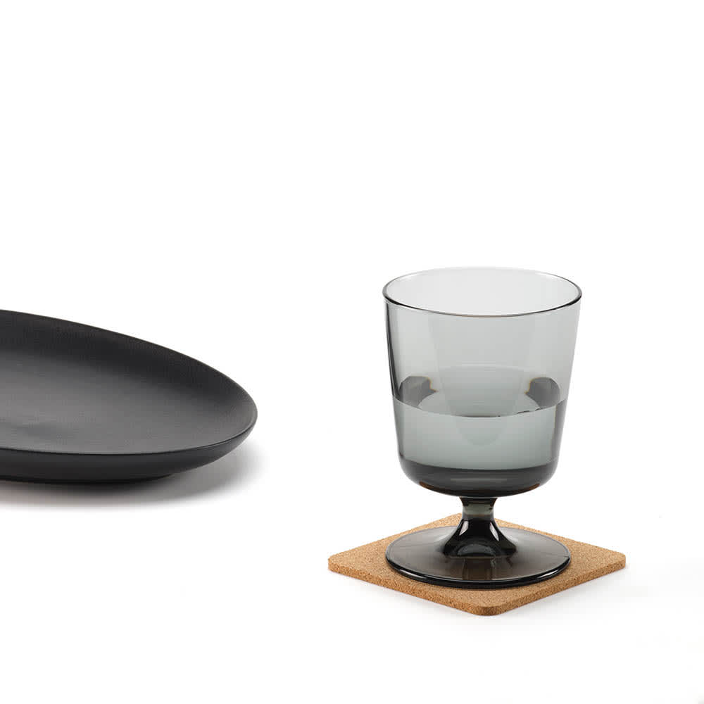 A cork coaster in a square shape supports a grey glass drink, accompanied by a black plate.