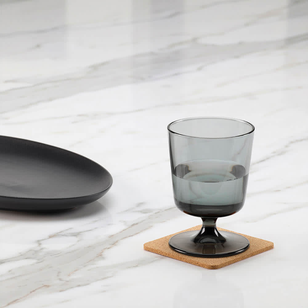 A grey drink glass sits on a square cork coaster on a marble surface, alongside a black plate.