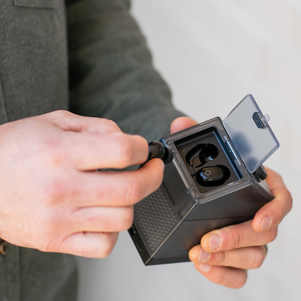 A person holding a compact black device with earbuds, featuring a transparent lid and a charging port.