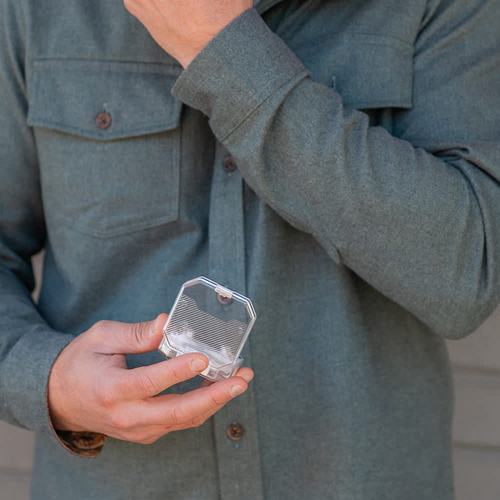 A person holding a clear, hexagonal product case, wearing a grey flannel shirt. The background is blurred.