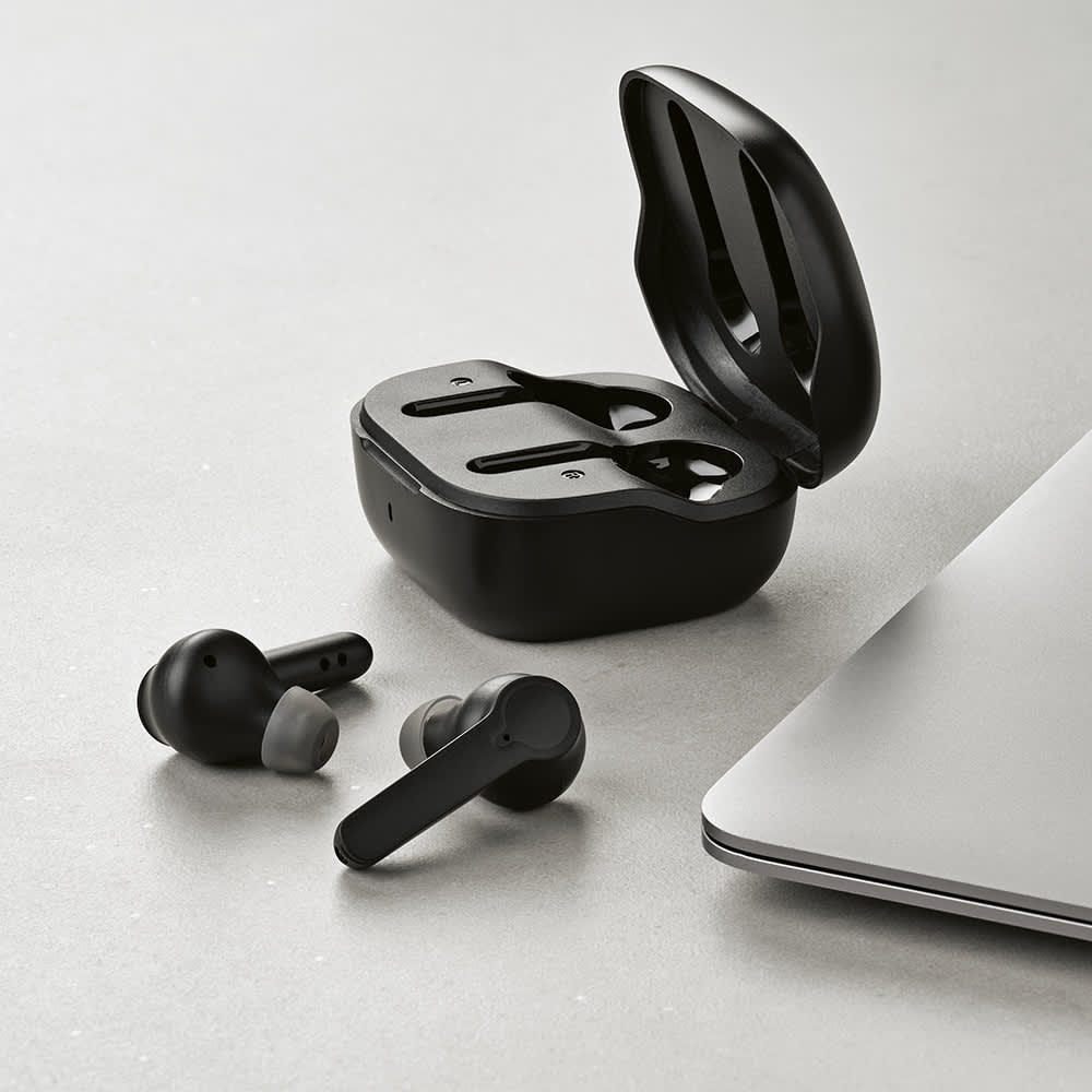 Black wireless earbuds in a charging case, placed next to a laptop on a light-coloured surface.