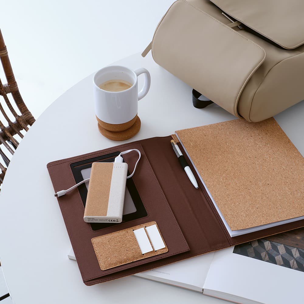 A cork A4 folder with a notepad, power bank, phone, and pen, set on a table with a cup of coffee and a bag.