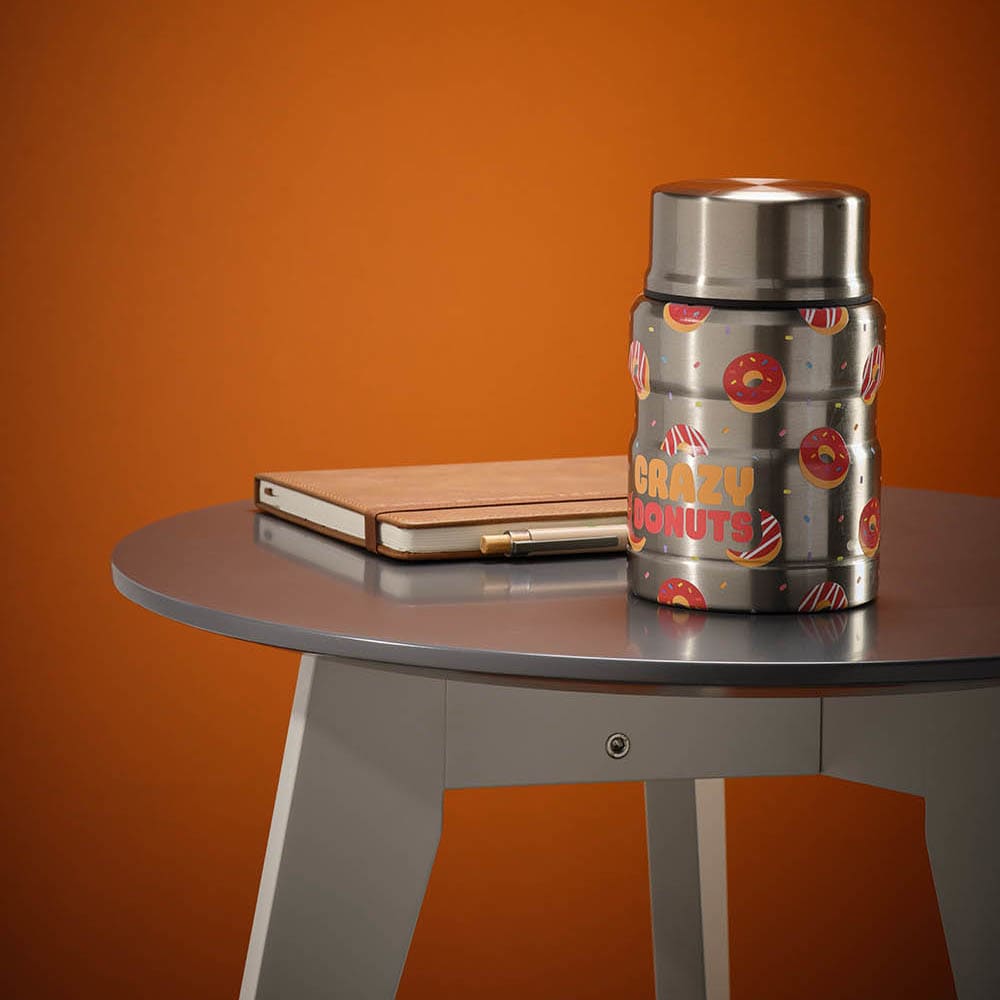 A drink bottle in silver with a fun donut print, placed on a table beside a closed laptop.