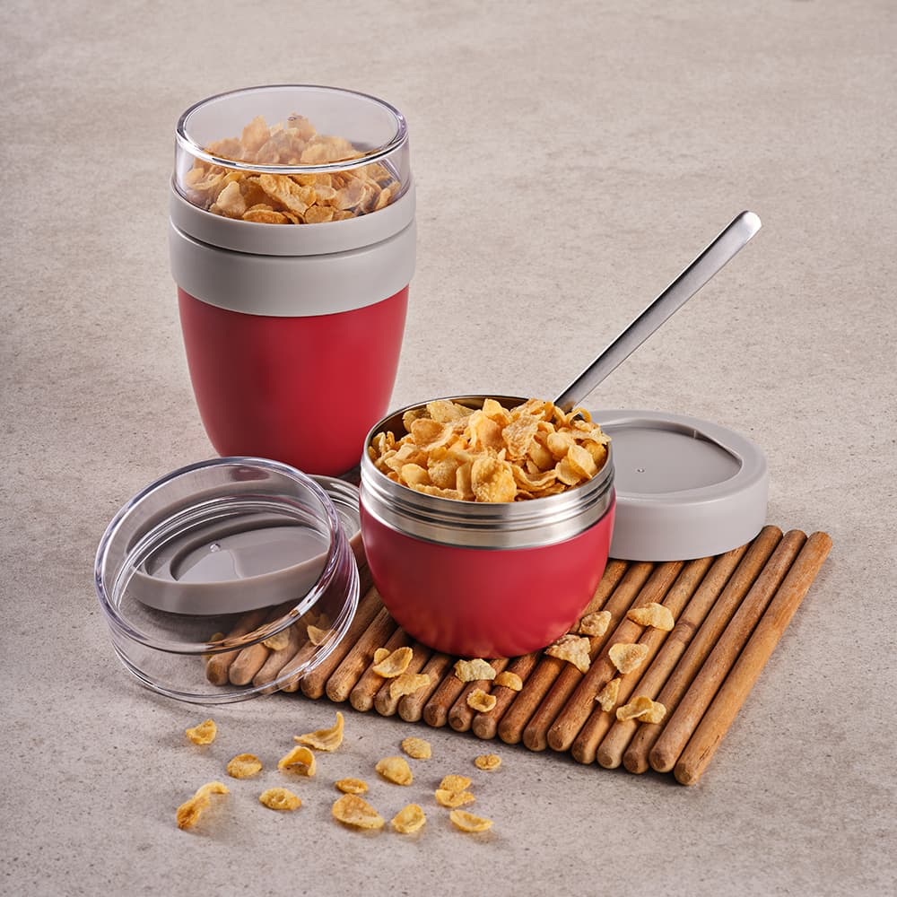 A red food flask with two compartments, a transparent lid, and a spoon, displayed on a wooden mat with cereal pieces.