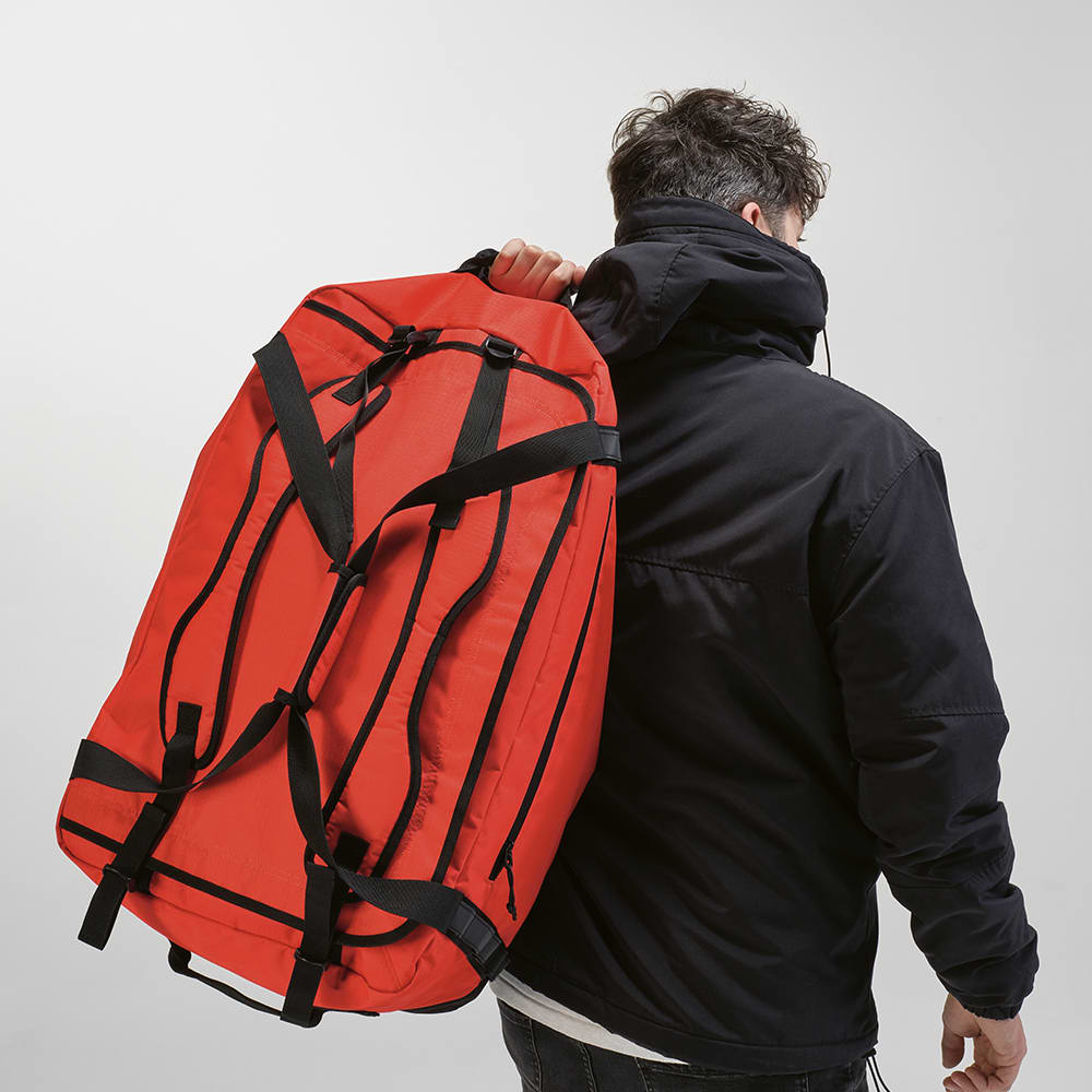 A large red gym bag with black straps being carried over the shoulder by a person in a black jacket.