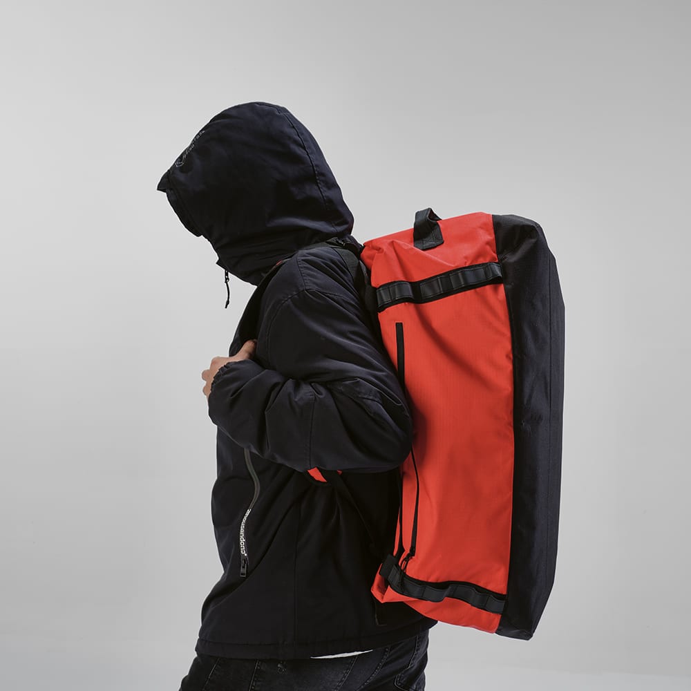 A person wearing a dark hooded jacket carries a large red and black gym bag on their back. The bag features reflective strips.