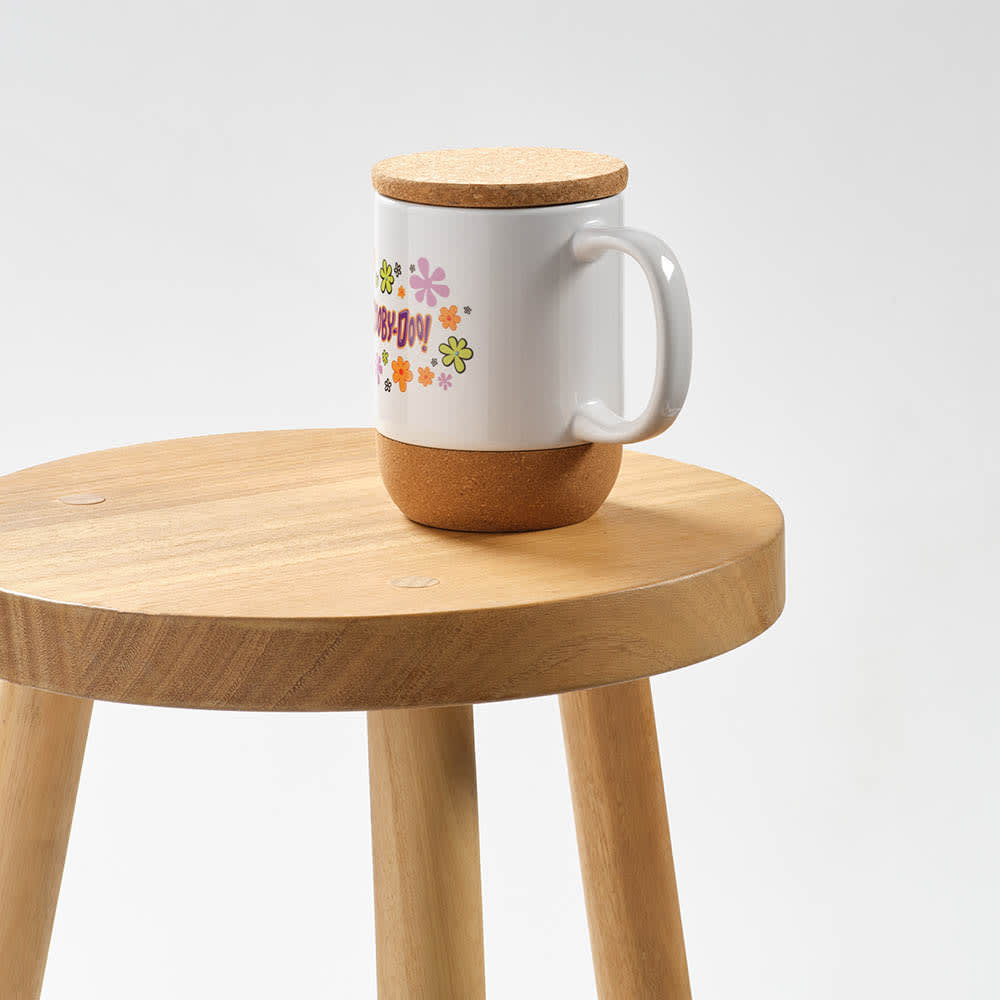 A ceramic mug with a cork lid and base, featuring a floral design, sits on a wooden stool.