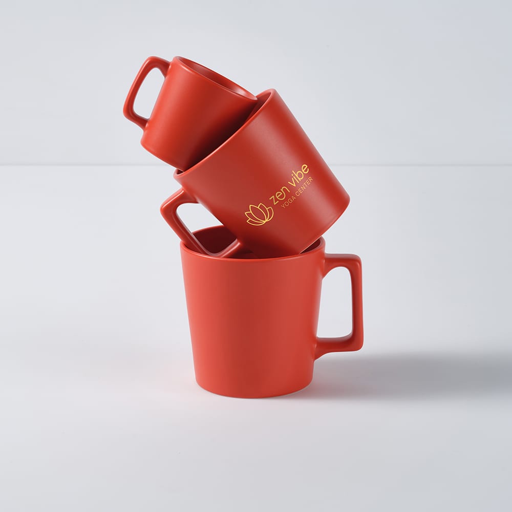 Three red ceramic mugs stacked together, featuring a small logo. Each mug has a matt finish.