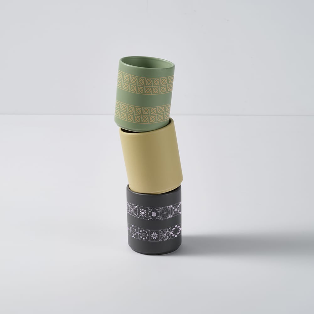 A stack of three ceramic mugs, featuring a matt finish in green, yellow, and dark grey with decorative patterns.